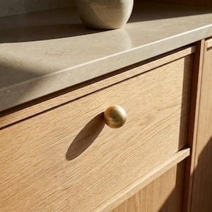 Op de afbeelding: Een ronde kastknop van geborsteld messing bevestigd aan een ladefront van licht eikenhout. De knop heeft een gladde, bolvormige vorm met een metalen afwerking en werpt een duidelijke schaduw over de houtnerf van het ladefront.
