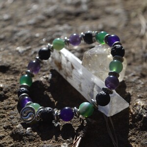 Może przedstawiać: Bransoletka wykonana z czarnych koralików z lawy wulkanicznej, koralików ametystowych, zielonych koralików agatowych i srebrnego charmsa z symbolem yin yang.