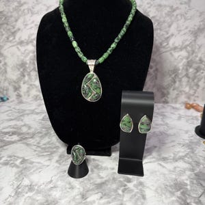 May include: A jewelry set featuring a necklace, a ring, and a pair of earrings. Each piece contains teardrop-shaped green and black stone inlays set in silver metal. The necklace has a beaded green and black stone chain with a large pendant.