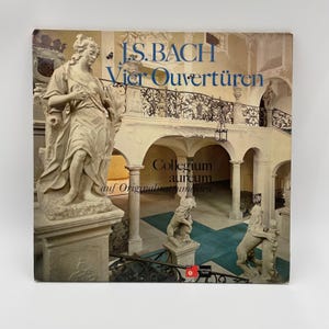 May include: A square vinyl record cover featuring a photograph of a grand interior with stone statues, a staircase, and dark metal railings. The text reads J.S. BACH Vier Ouvertüren, Collegium aureum auf Originalinstrumenten, and Harmonia mundi.