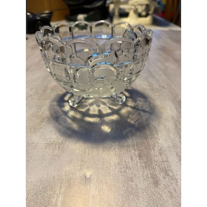 May include: A clear glass bowl featuring a scalloped rim and three small feet. The exterior surface is decorated with a repeating frosted floral pattern that contrasts with the transparent glass, creating a textured and detailed appearance.