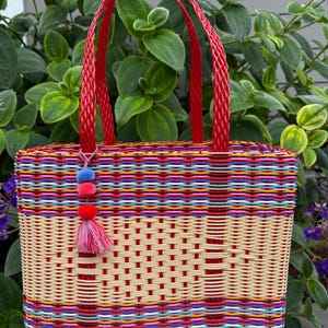 May include: A woven tote bag with a red handle and a colorful striped pattern. The bag features a beige center section with a grid design and a tassel with pom-poms. The bag is made of woven plastic.