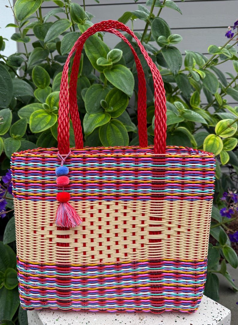 May include: A woven tote bag with a red handle and a colorful striped pattern. The bag features a beige center section with a grid design and a tassel with pom-poms. The bag is made of woven plastic.