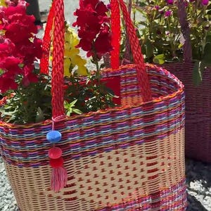 May include: Woven basket with red handles, filled with red and yellow flowers. The basket has a colorful woven pattern with pink, blue, and orange stripes. A decorative tassel hangs from the side. Another basket with purple handles is in the background.