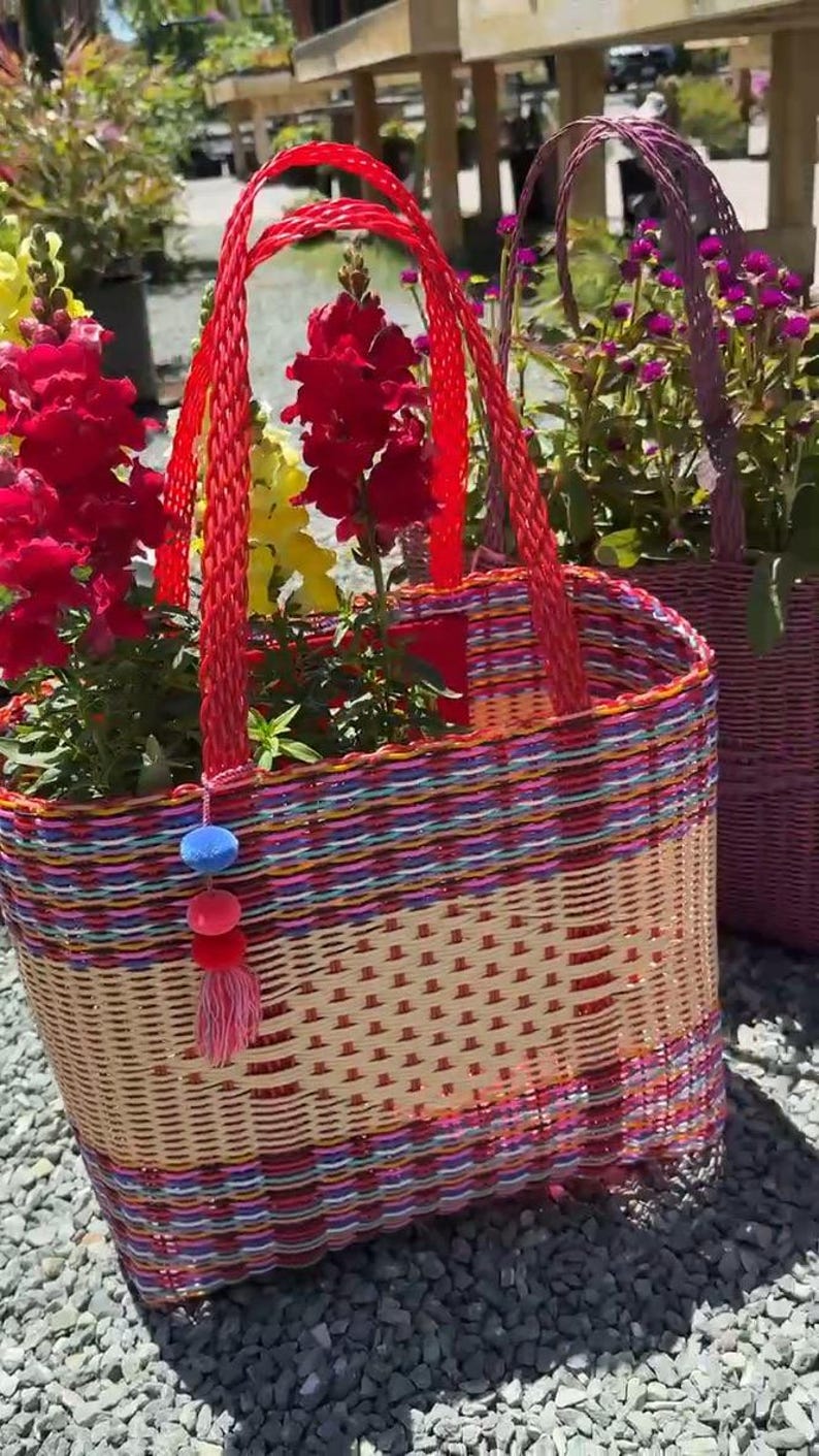 May include: Woven basket with red handles, filled with red and yellow flowers. The basket has a colorful woven pattern with pink, blue, and orange stripes. A decorative tassel hangs from the side. Another basket with purple handles is in the background.