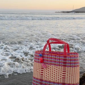 May include: A woven tote bag with red handles and colorful stripes. The bag is decorated with a tassel and pom-poms. The bag is sitting on a rock near the ocean. The bag is approximately 12 inches wide and 10 inches tall.