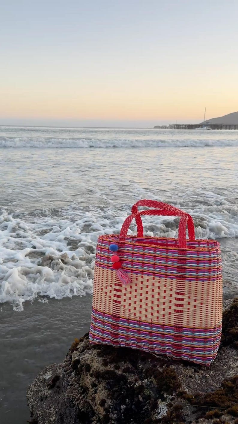 May include: A woven tote bag with red handles and colorful stripes. The bag is decorated with a tassel and pom-poms. The bag is sitting on a rock near the ocean. The bag is approximately 12 inches wide and 10 inches tall.