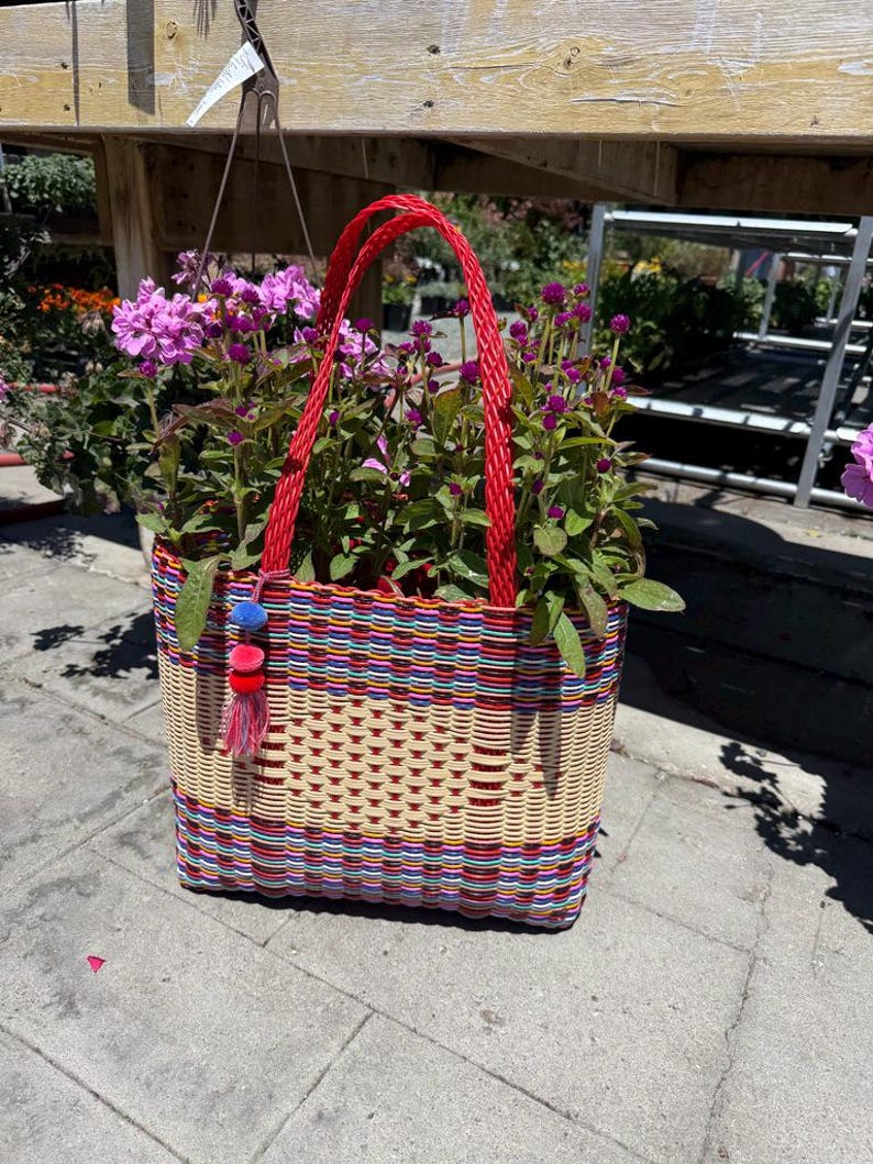 May include: A woven basket with a red handle and colorful stripes, filled with vibrant purple flowers. The basket has a tassel with blue, red, and yellow accents. The basket is sitting on a gray surface.