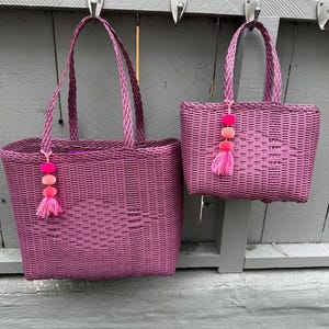 May include: Two woven magenta tote bags with matching handles and decorative pink pom-poms. The bags are rectangular with a woven pattern and are hanging from metal hooks against a gray wooden background. The bags are different sizes.