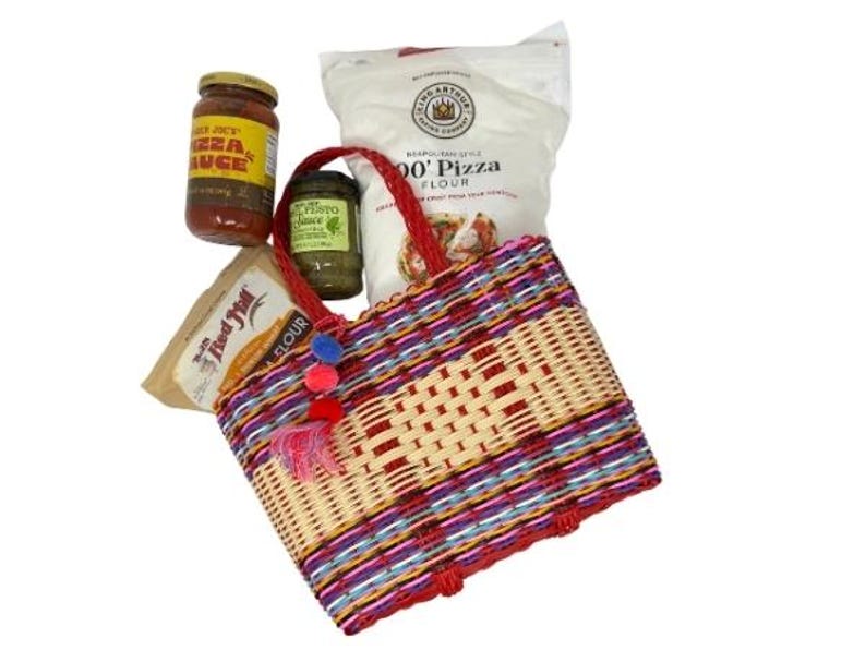 May include: A woven tote bag with red handles, filled with pizza-making ingredients. The bag features a colorful pattern of red, purple, and blue stripes. Jars of pizza sauce and pesto, along with bags of flour, are visible.