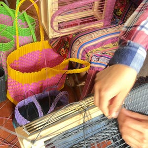 May include: A collection of woven baskets in various stages of completion. The baskets are in shades of yellow, pink, green, and purple. A wooden frame is being used to weave a basket with pink material.