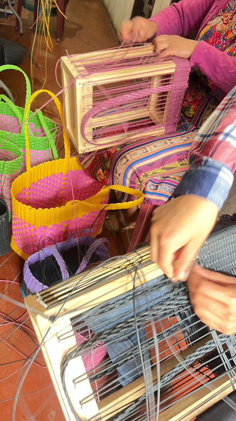 May include: A collection of woven baskets in various stages of completion. The baskets are in shades of yellow, pink, green, and purple. A wooden frame is being used to weave a basket with pink material.