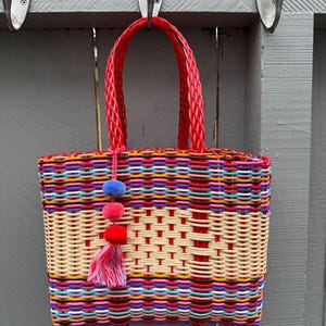 May include: A colorful woven tote bag with a red handle and a decorative tassel. The bag features a vibrant mix of colors, including red, yellow, blue, and pink. The bag is hanging on a gray wooden fence.
