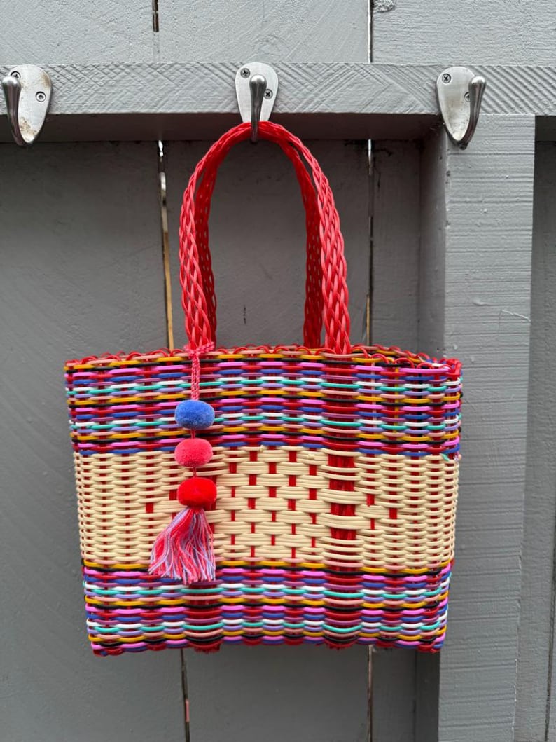May include: A colorful woven tote bag with a red handle and a decorative tassel. The bag features a vibrant mix of colors, including red, yellow, blue, and pink. The bag is hanging on a gray wooden fence.