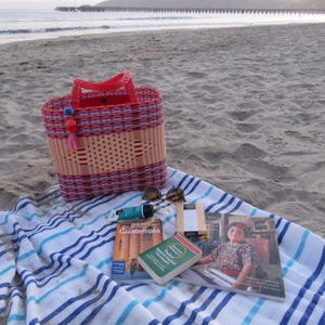 May include: A woven beach bag with red handles and a tassel, resting on a striped beach towel. Sunglasses, books about Guatemala, and a tube of sunscreen are also on the towel, with a sandy beach and ocean in the background.