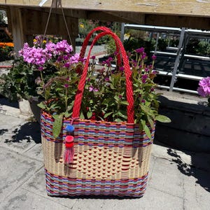 May include: A woven basket with red handles, filled with purple flowers. The basket is made of colorful, interwoven plastic strips in red, blue, and yellow. A small tassel with blue, red, and pink accents hangs from the side.