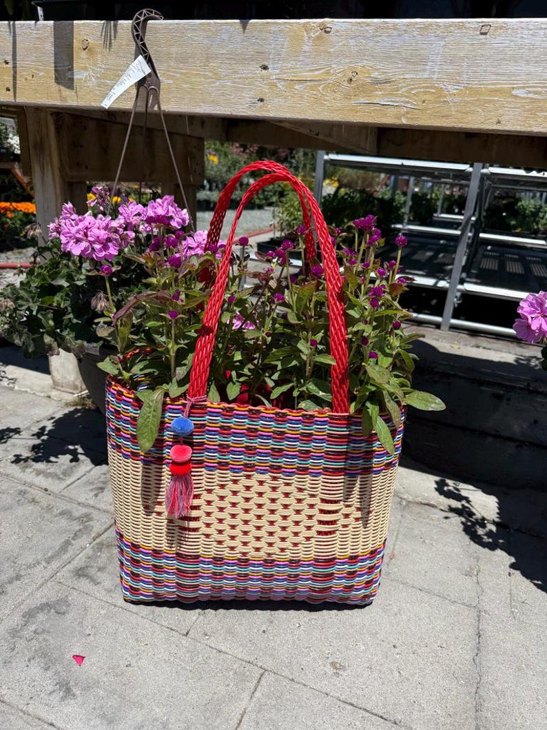 May include: A woven basket with red handles, filled with purple flowers. The basket is made of colorful, interwoven plastic strips in red, blue, and yellow. A small tassel with blue, red, and pink accents hangs from the side.