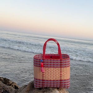 May include: A rectangular woven tote bag with a red handle and trim. The bag features a woven pattern in natural, red, and purple hues. A tassel and pom-pom accent hangs from the side. The bag is sitting on a rock near the ocean.
