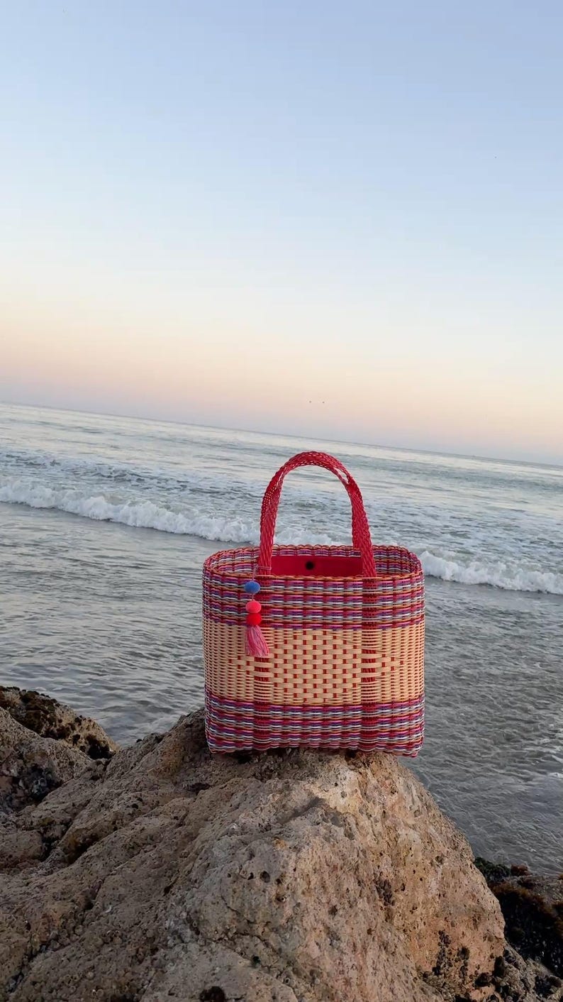 May include: A rectangular woven tote bag with a red handle and trim. The bag features a woven pattern in natural, red, and purple hues. A tassel and pom-pom accent hangs from the side. The bag is sitting on a rock near the ocean.