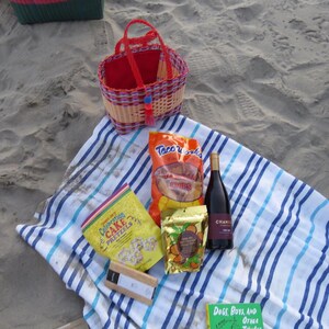May include: A picnic scene on a sandy beach. A woven basket with a red interior and handle sits atop a striped towel. Snacks include chips, pretzels, and nuts, along with a bottle of wine and a book titled "Dogs, Boys, and Other Things".