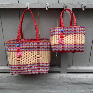 May include: Two woven tote bags with red handles and colorful woven patterns. The bags are rectangular with a light beige center and are decorated with a tassel of pink and blue pom-poms. The bags are hanging on a gray wooden surface.