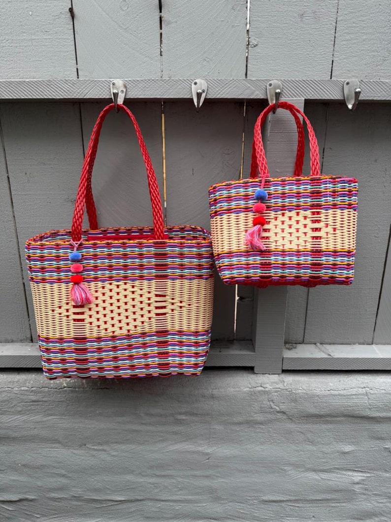 May include: Two woven tote bags with red handles and colorful woven patterns. The bags are rectangular with a light beige center and are decorated with a tassel of pink and blue pom-poms. The bags are hanging on a gray wooden surface.