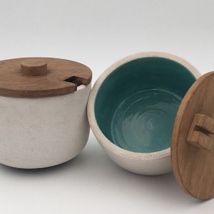 Puede incluir: Dos cuencos de cerámica con tapas de madera. Un cuenco es blanco con un interior turquesa, el otro es blanco con un interior blanco. Ambas tapas tienen un pomo en la parte superior.