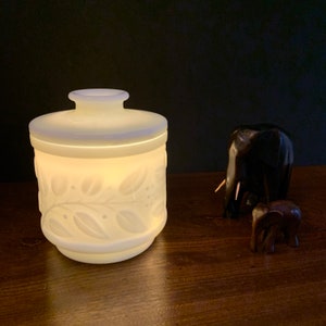 Puede incluir: Jarrón de cerámica blanca con un diseño floral. El jarrón tiene una tapa y está iluminado desde el interior. Dos figuritas de elefante de madera están en el fondo.