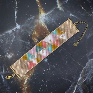 Puede incluir: Un brazalete de cuentas de colores con un patrón geométrico en tonos de rosa, marrón, amarillo y turquesa. El brazalete tiene un cierre y una cadena dorados.