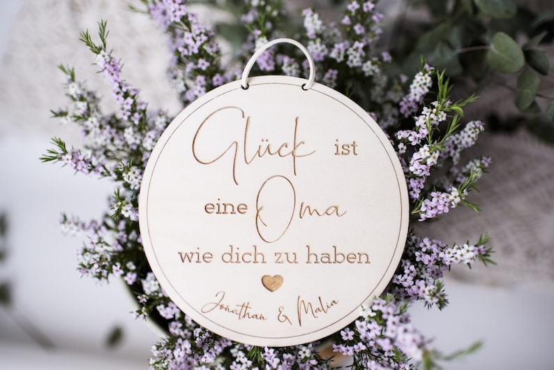 Puede incluir: Un letrero redondo de madera blanca con el texto "Gl&uuml;ck ist eine Oma wie dich zu haben" y los nombres "Jonathan & Malia" escritos en cursiva. El letrero est&aacute; colgado de una cuerda blanca y est&aacute; rodeado de flores moradas.