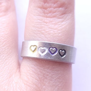 May include: A silver ring with five hearts in a row. The hearts are yellow, white, purple, white, and yellow.