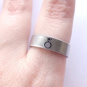 以下が含まれることがあります： 星が刻まれたジェンダーニュートラルなシンボルが刻まれた銀の指輪。シンボルは星が内側に刻まれた円です。