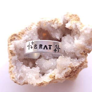 Peut inclure: Une bague en argent avec le mot "BRAT" gravé dessus, entourée de petites croix. La bague repose sur un cristal blanc et brun.