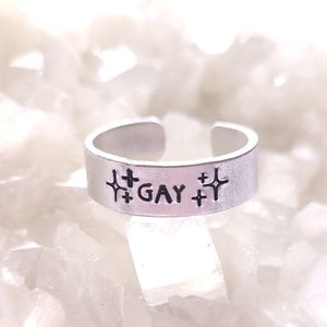 May include: A silver adjustable ring with the word "GAY" and stars on either side.