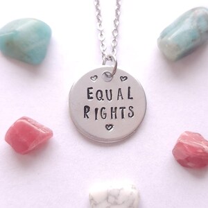 Peut inclure: Un collier avec un pendentif circulaire argenté portant l'inscription gravée EQUAL RIGHTS accompagnée de petits cœurs au-dessus et en dessous des mots. Le pendentif est suspendu à une fine chaîne argentée et entouré de cinq pierres polies.