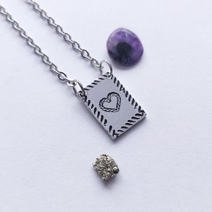 May include: A silver necklace with a heart-shaped charm on a rectangular tag. The tag has a rope-like border. The necklace also includes a purple stone and a small, rough, gold-colored stone.