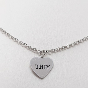 Peut inclure: Un collier en chaîne argent avec un pendentif en forme de cœur argenté sur lequel est inscrit "THEY" en lettres noires.