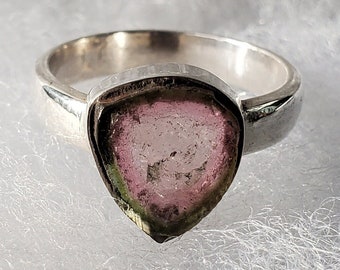 Può includere: Un anello d'argento con una pietra preziosa di tormalina multicolore a forma di pera. La pietra preziosa mostra sfumature di rosa, verde e trasparente, incastonata in una fascia d'argento lucida. L'anello è esposto su una superficie bianca strutturata.