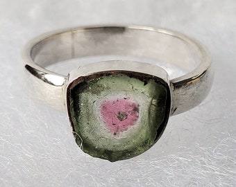 Può includere: Un anello d'argento con una pietra preziosa verde, rosa e bianca. La pietra preziosa è di forma quadrata e presenta un motivo unico.