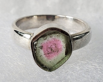 Può includere: Un anello d'argento con una pietra preziosa rosa, verde e bianca. La pietra preziosa ha una forma esagonale e un motivo unico.