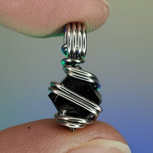 May include: A black gemstone pendant wrapped in silver wire. The pendant is shaped like a square with rounded edges and has a shiny, metallic finish.