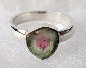 Könnte beinhalten: Ein Silberring mit einem dreieckigen Wassermelonen-Turmalin-Edelstein. Der Edelstein zeigt einen Farbverlauf mit einem rosa Zentrum, das in Grün übergeht, und ist in einem polierten Silberband gefasst. Der Ring wird auf einer weißen, strukturierten Oberfläche präsentiert.