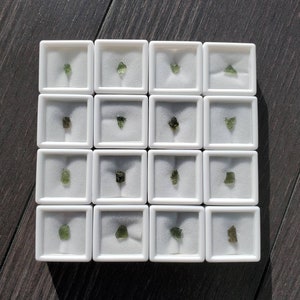 Op de afbeelding: Zestien kleine, onregelmatig gevormde, groene edelstenen worden gepresenteerd in individuele witte vierkante doosjes. De doosjes zijn gerangschikt in een rasterpatroon op een donker houten oppervlak, met elke edelsteen gecentreerd.