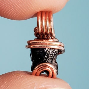 May include: A black gemstone pendant wrapped in copper wire. The gemstone is a dark, almost black, color and has a rough, natural texture. The copper wire is wrapped around the gemstone in a decorative pattern.