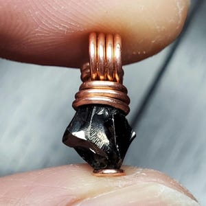 May include: A black obsidian stone pendant wrapped in copper wire. The stone is rough and has a natural, irregular shape. The copper wire is wrapped around the stone in a spiral pattern, creating a unique and eye-catching design.