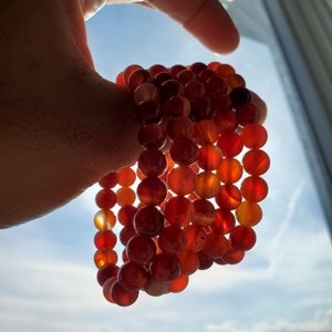 May include: A stack of red carnelian gemstone bracelets. The bracelets are made of round beads and are strung together on a clear elastic cord.