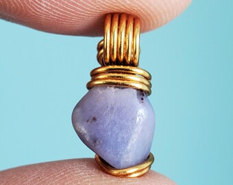 May include: A small, light purple gemstone pendant with gold wire wrapping. The gold wire forms a loop at the top and wraps around the stone. The pendant is held between two fingers against a light blue background.