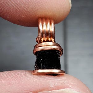 May include: A black gemstone pendant wrapped in copper wire. The pendant is a dark, opaque stone with a rough, natural texture. The copper wire is wrapped around the stone in a series of tight coils, creating a decorative and secure setting.