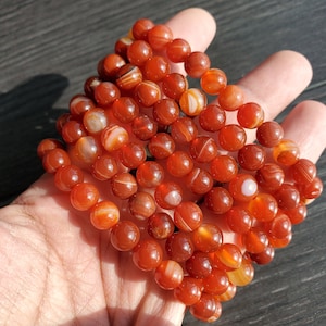 May include: Multiple strands of carnelian gemstone beads, each strand is made of round, smooth, orange-red beads with white stripes.