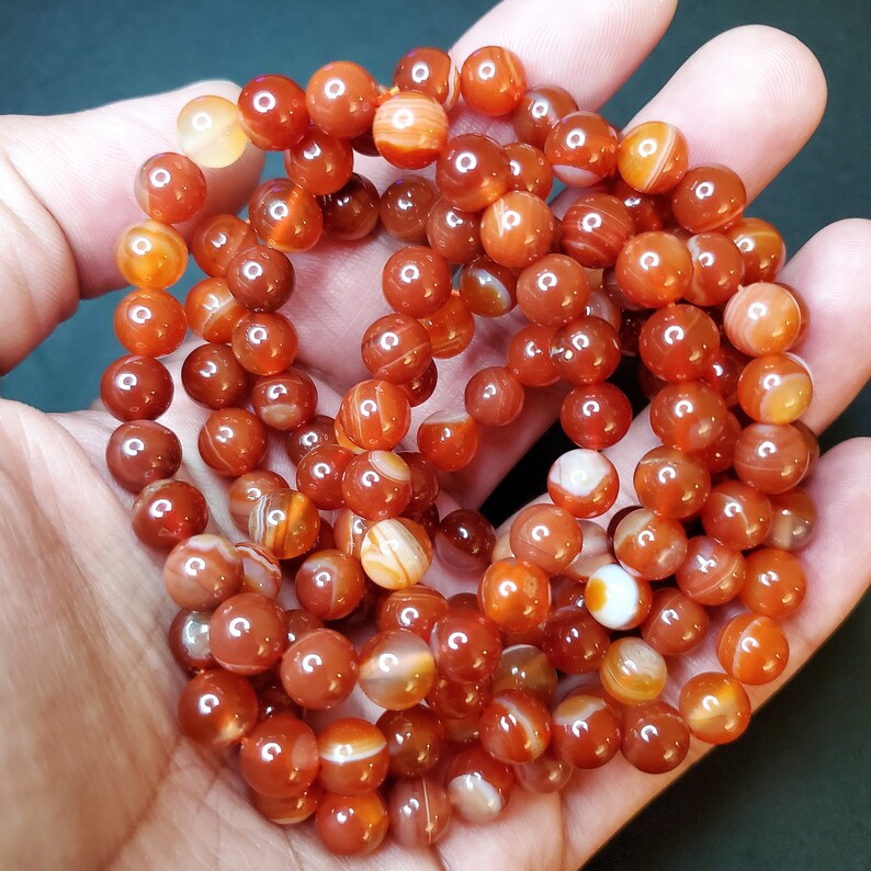 May include: A strand of carnelian beads, featuring a mix of red and orange tones with white banding. The beads are round and smooth, with a polished finish.